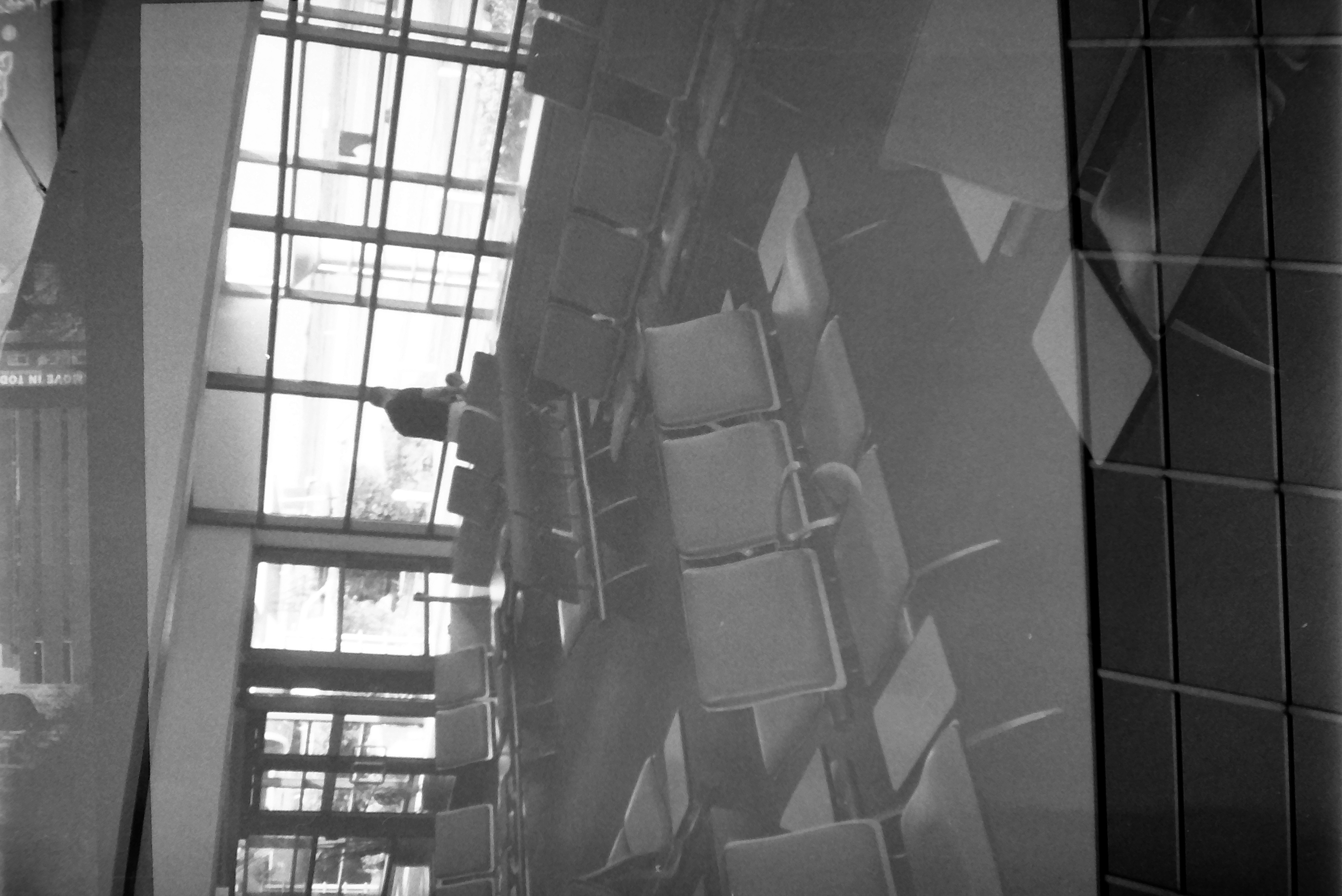 Rows of empty seats face a wall of gridded windows where one person stands, while a dark grid of panes overlays the lower floor.