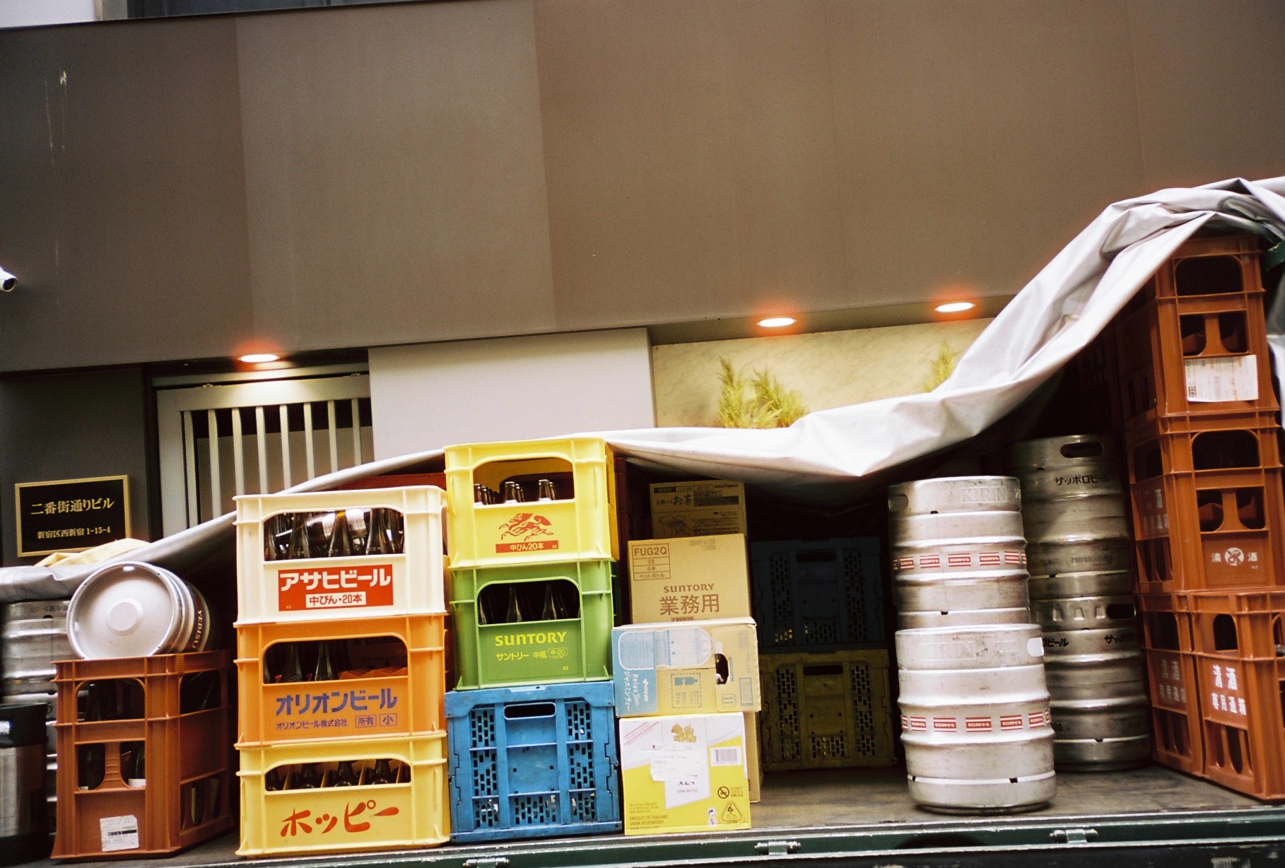 An open truck bed holds plastic crates labeled Asahi, Orion, and Hoppy beer beside Suntory boxes and steel kegs under a tarp.