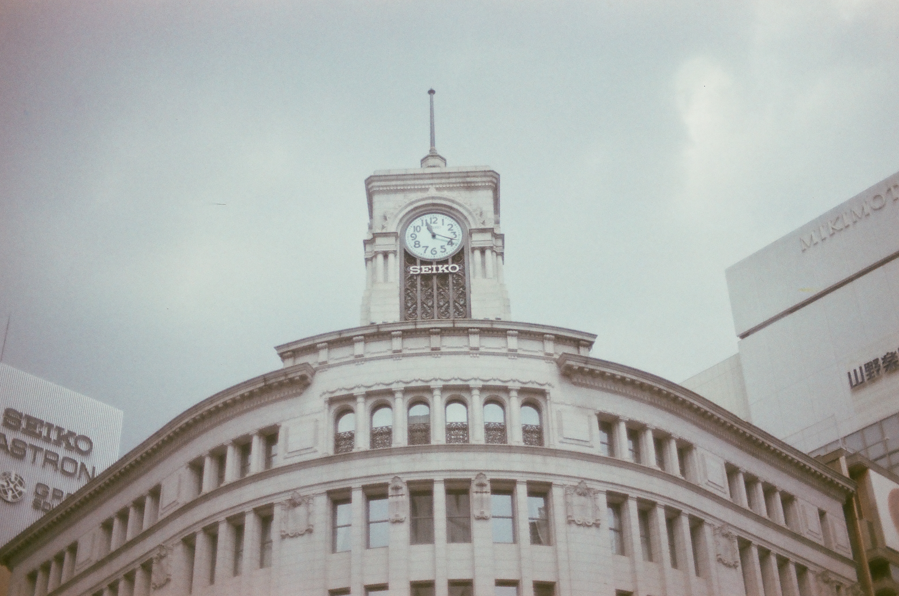 A white clock tower lettered 'SEIKO' below its dial crowns a curved stone building with rows of arched windows under an overcast sky.