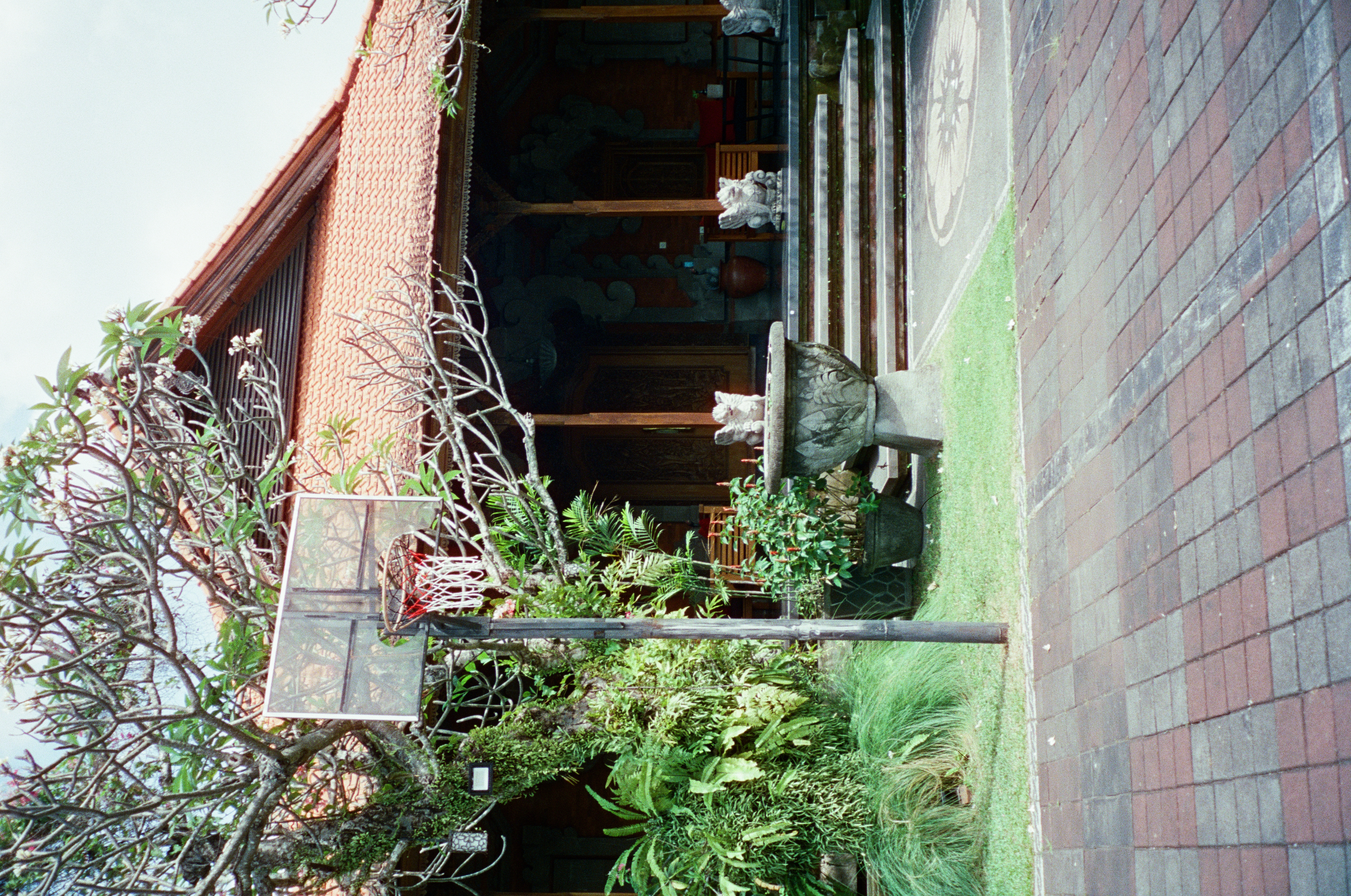 A basketball hoop on a dark pole stands on brick paving in front of an open-sided pavilion with a red tile roof, stone figures, and potted plants.