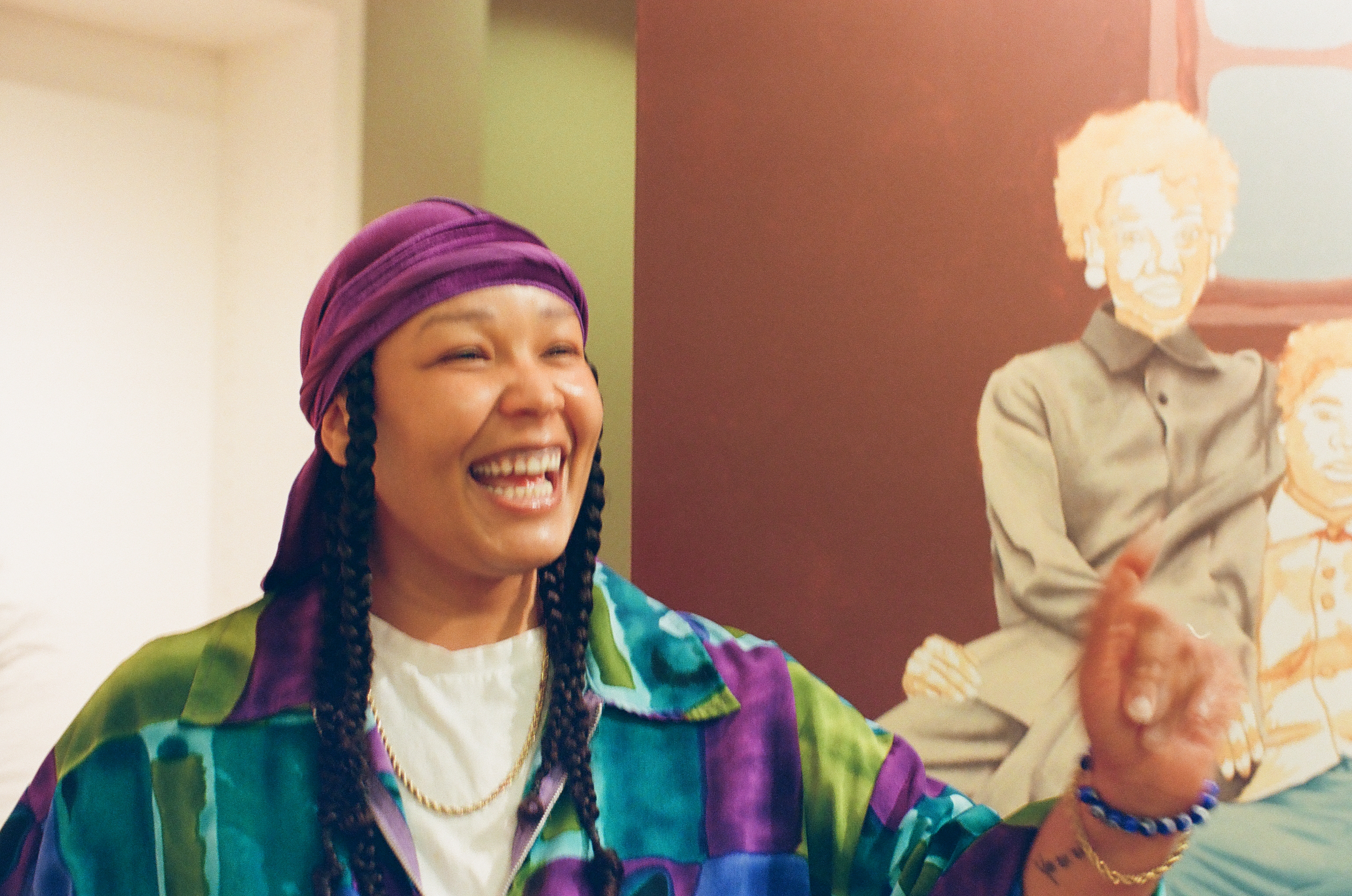 A laughing person gestures in bright light beside a large canvas painting of seated figures.