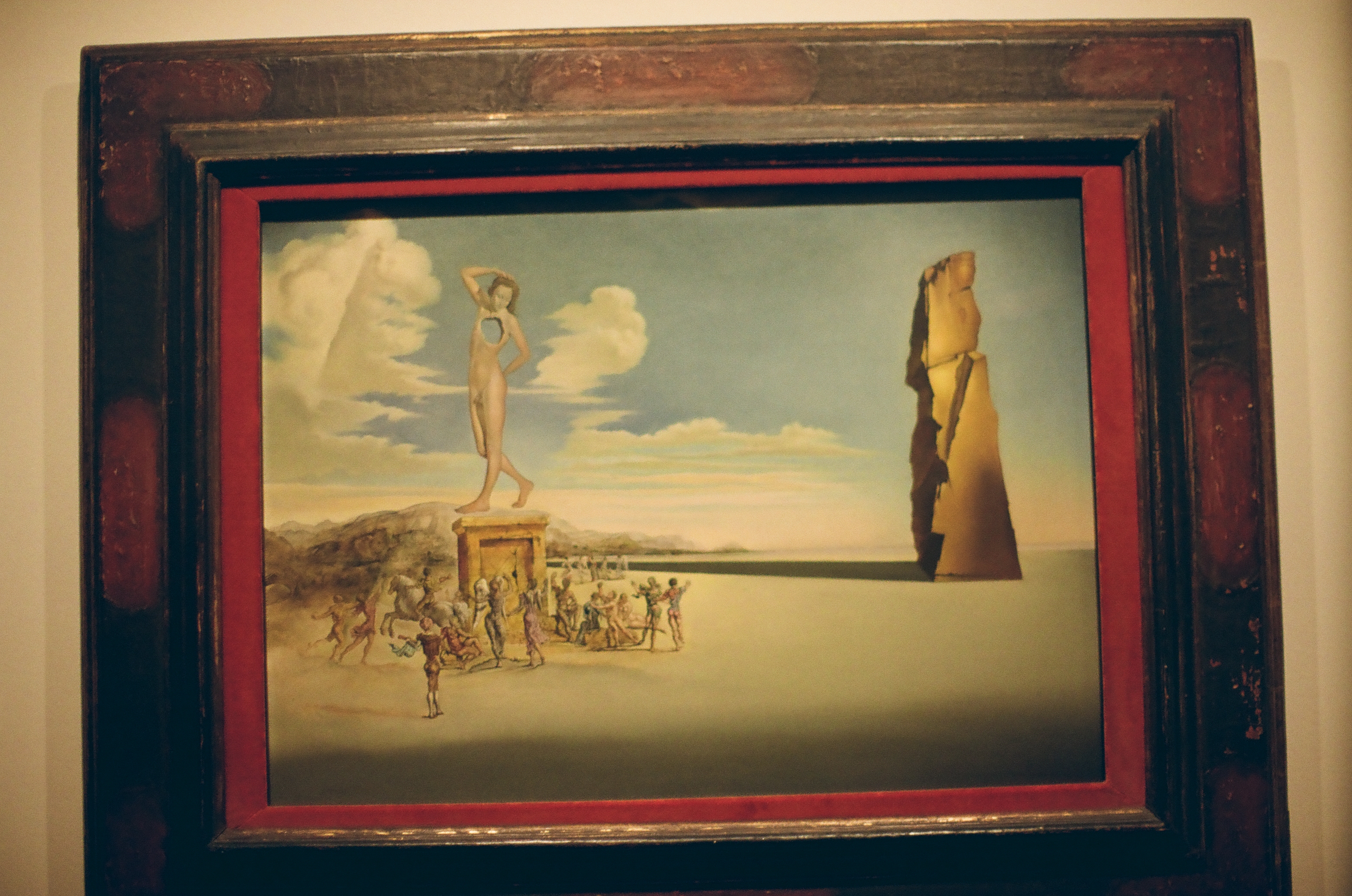 A dark wooden frame with a red inner border holds a painting of a nude figure on a pedestal above a crowd of small figures.
