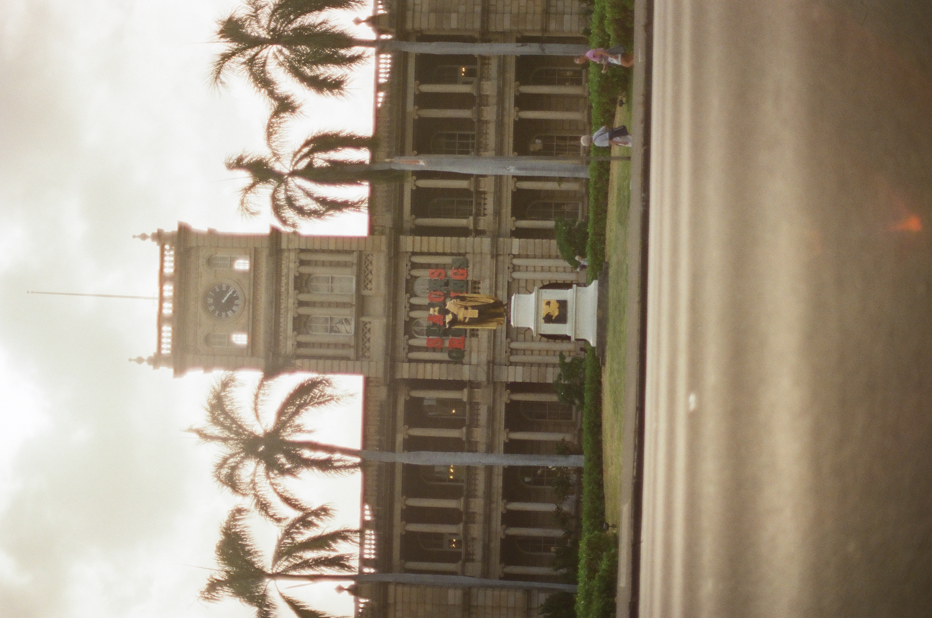 A pale symmetrical building with a central tower stands behind palms across a broad empty road.