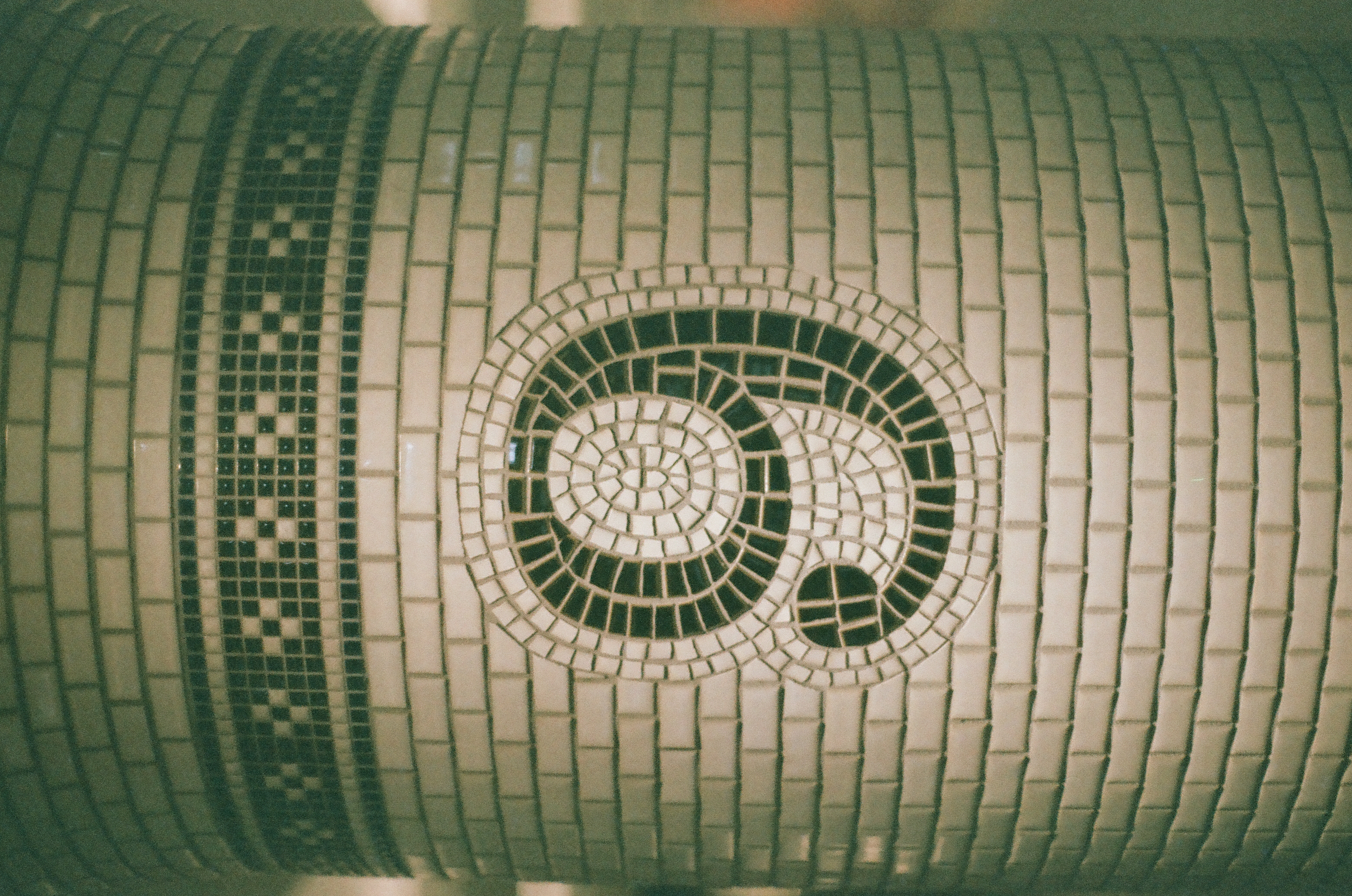 A tight vertical view of a tiled column shows the numeral nine formed from dark mosaic tesserae in a spiral shape beneath a checkered band.