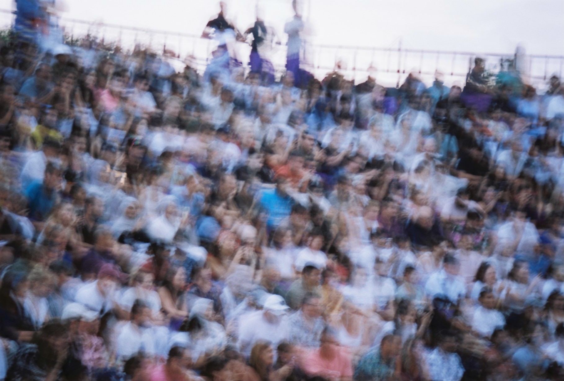A densely packed crowd blurs across tiered seating, with several standing figures along a railing beneath a pale sky.