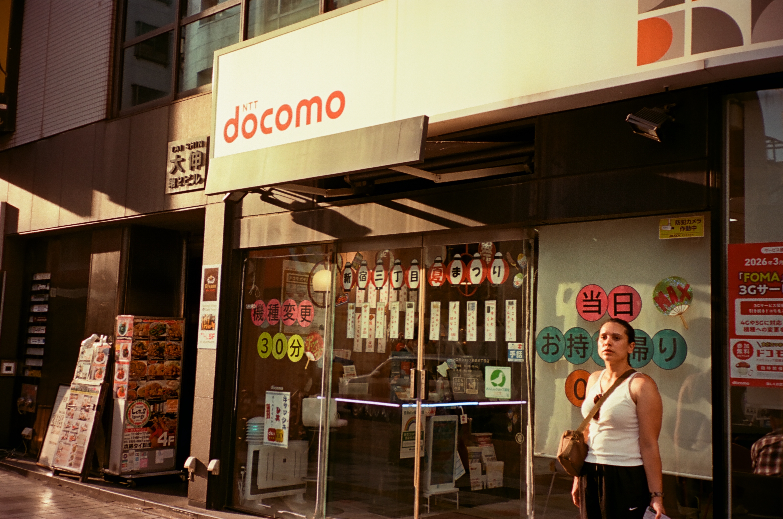 A woman in a white tank top with a crossbody bag stands outside a shop whose sign reads docomo, with hanging paper signs in the window.