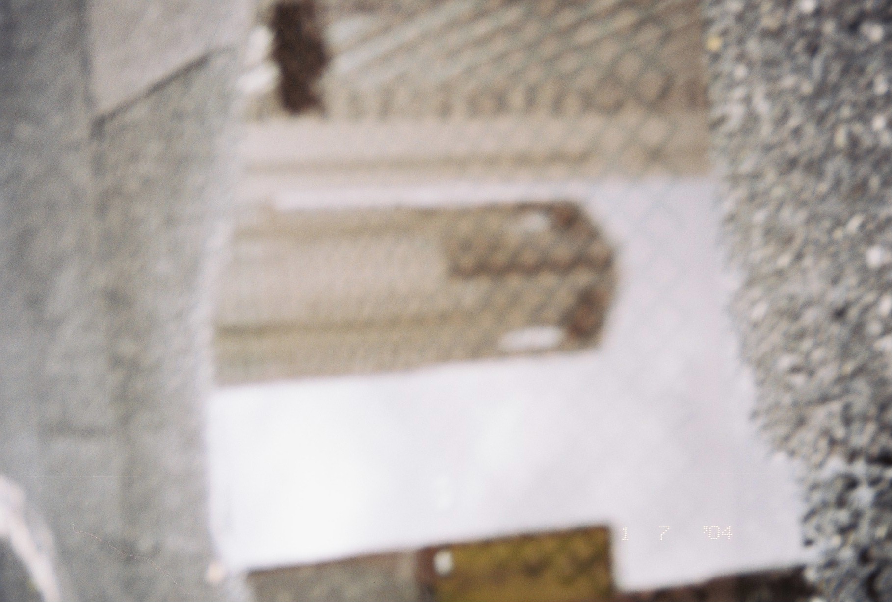 A soft-focus puddle on gravelly pavement reflects a pale gridded tower, a white sky, and a yellow rectangle at the upper right.