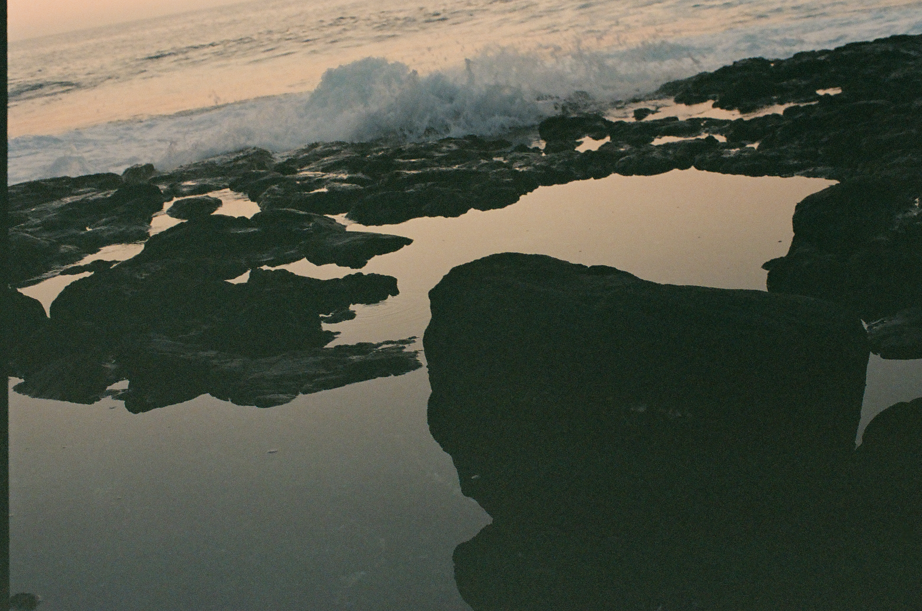 Still tide pools reflect a pale peach sky among broad dark rocks while surf breaks beyond.