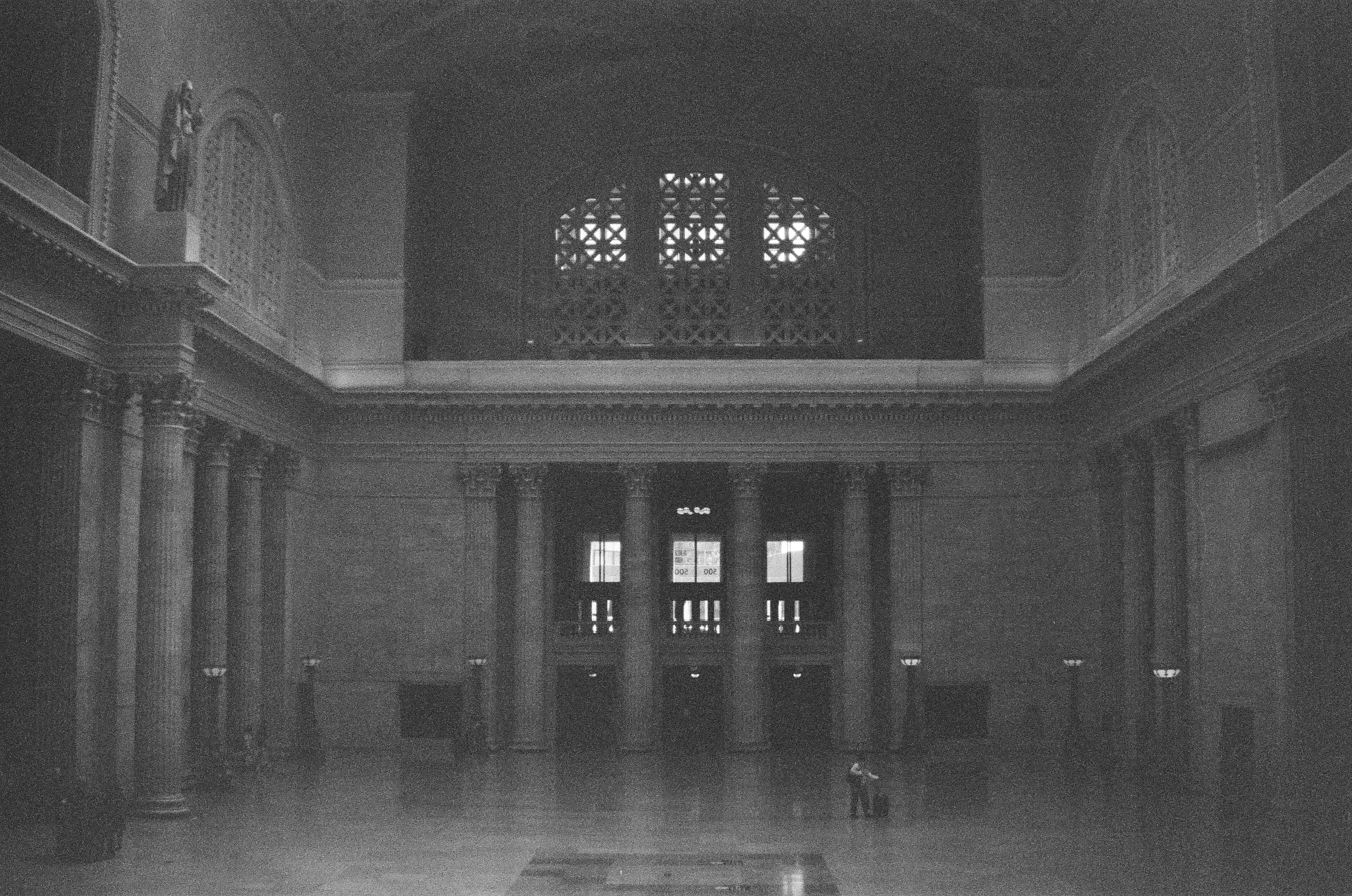 Tall columns and a distant doorway face a wide polished floor beneath small patterned windows.