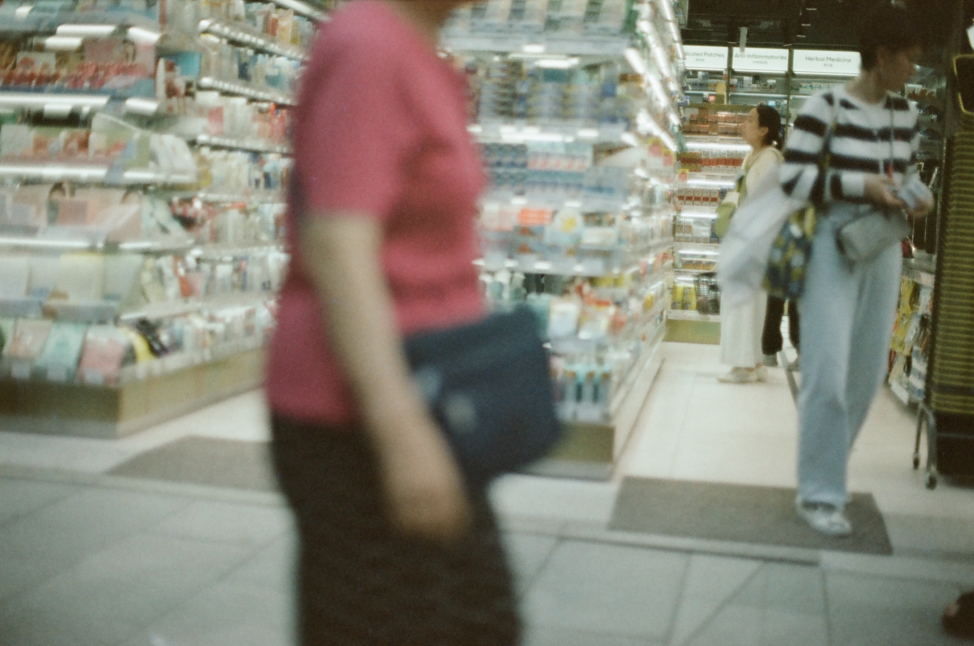 A person in a red shirt blurs across a drugstore aisle foreground, with shelves labeled 'Herbal Medicine' and 'Anti-Inflammatories' behind two shoppers.