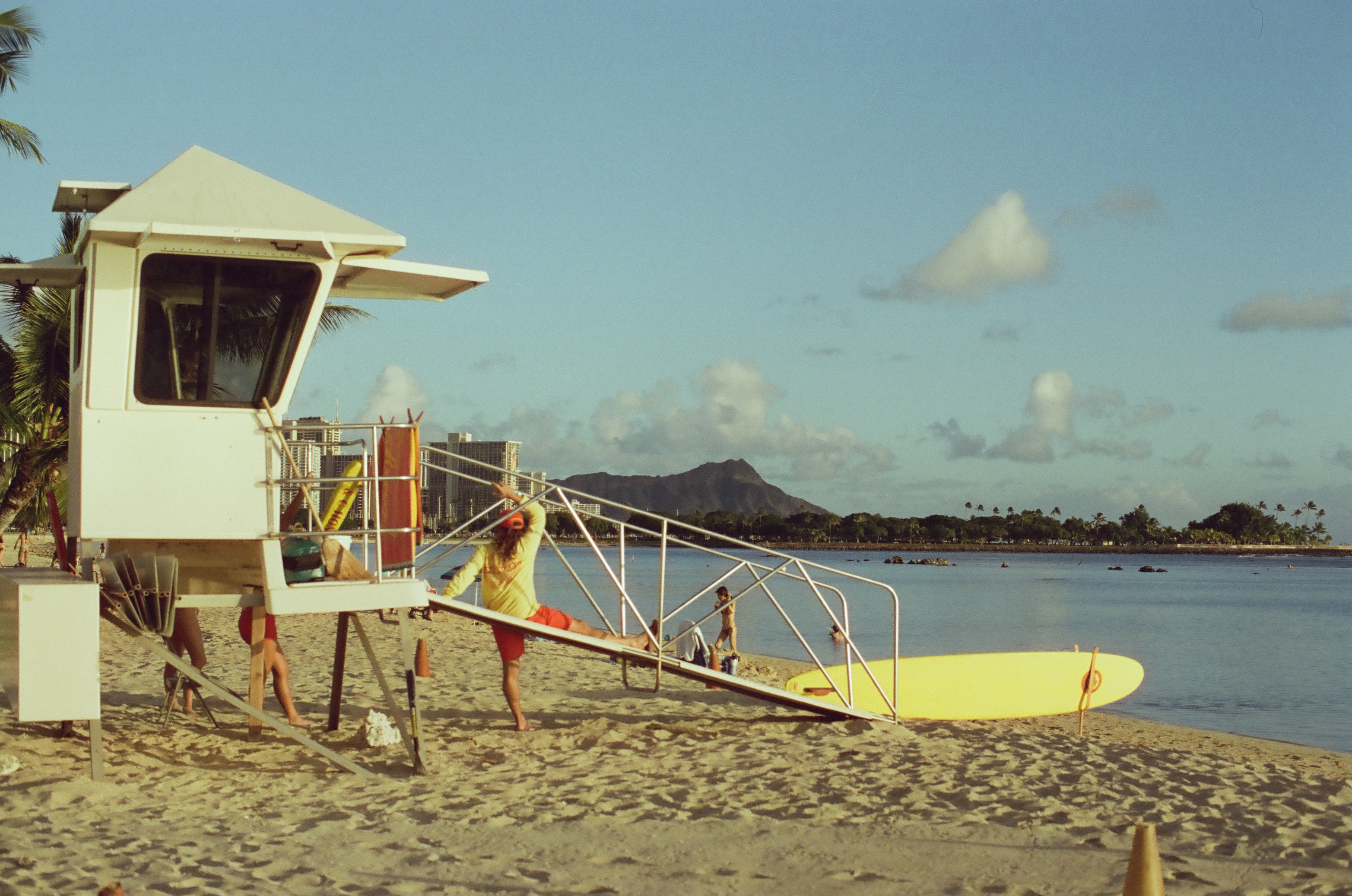 A lifeguard in a yellow shirt and red shorts stretches one leg up the tower's ramp rail, the peaked mountain rising sharply behind the shoreline.