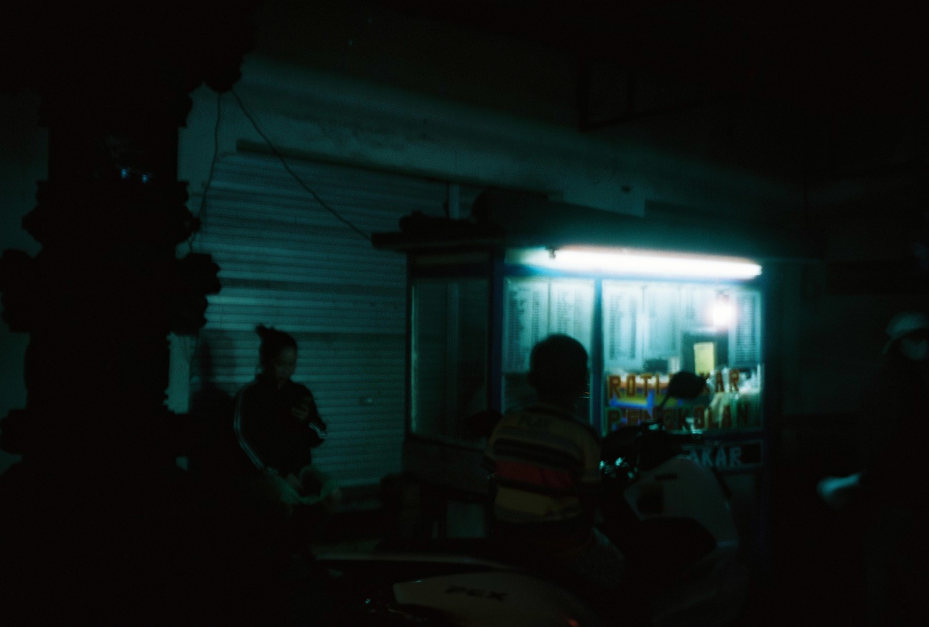 Silhouetted people and two-wheeled vehicles frame a lit opening where “ROTI” and “BAKAR” are readable; other text is obscured or cropped.