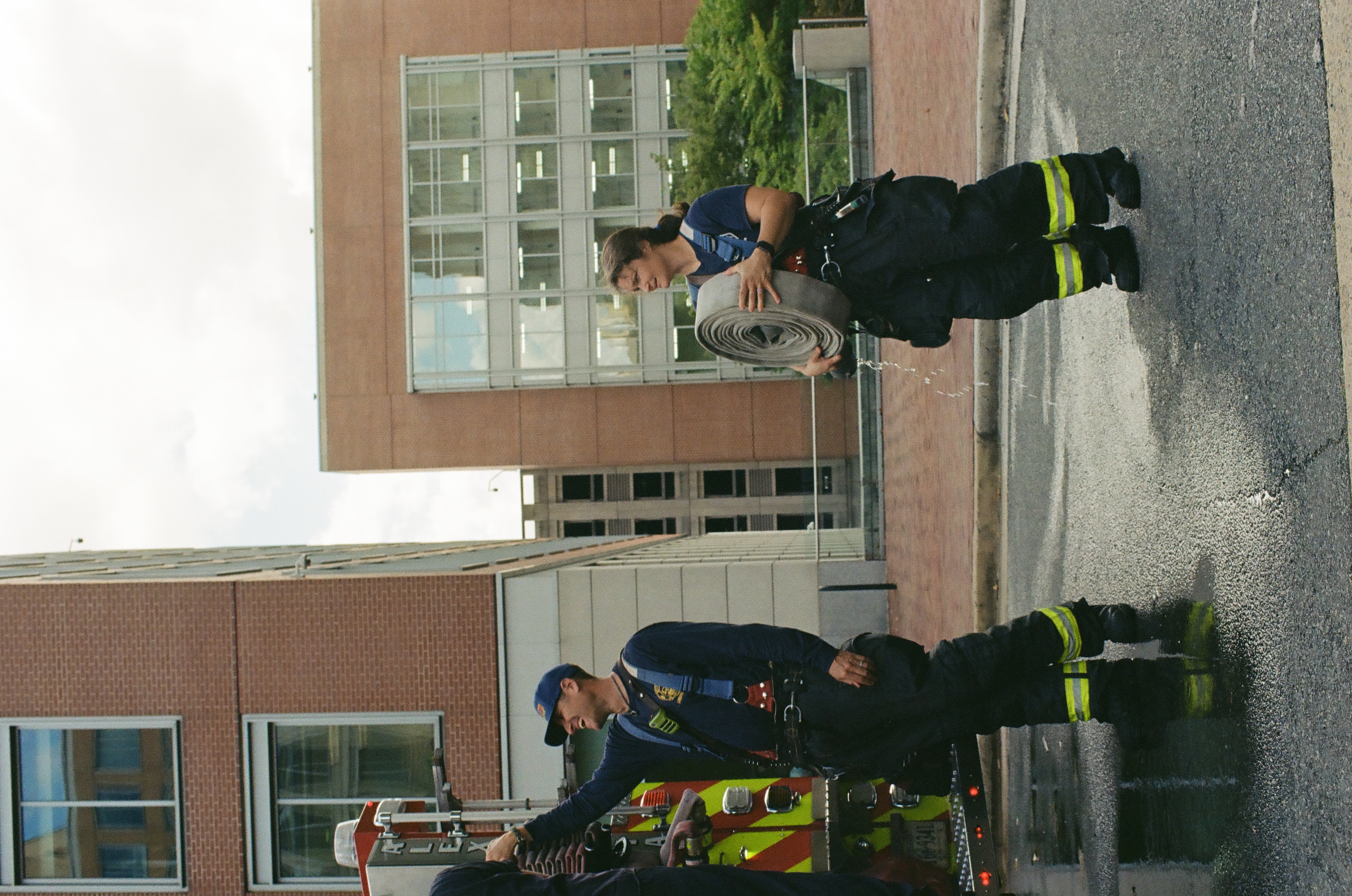 Two firefighters in dark turnout gear stand beside a fire engine, one carrying a dripping coiled hose on a wet street.