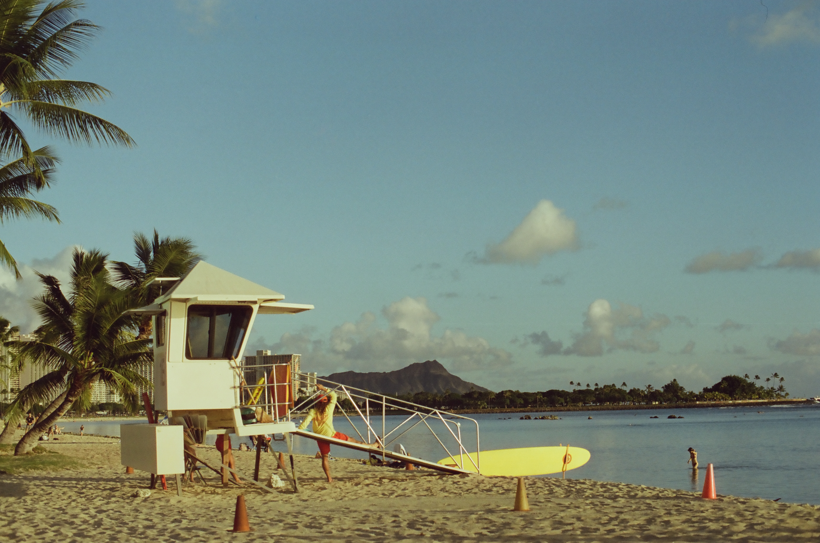 A white lifeguard tower stands on the sand beside a yellow paddleboard, its ramp railing draped with red gear as someone stretches on it.